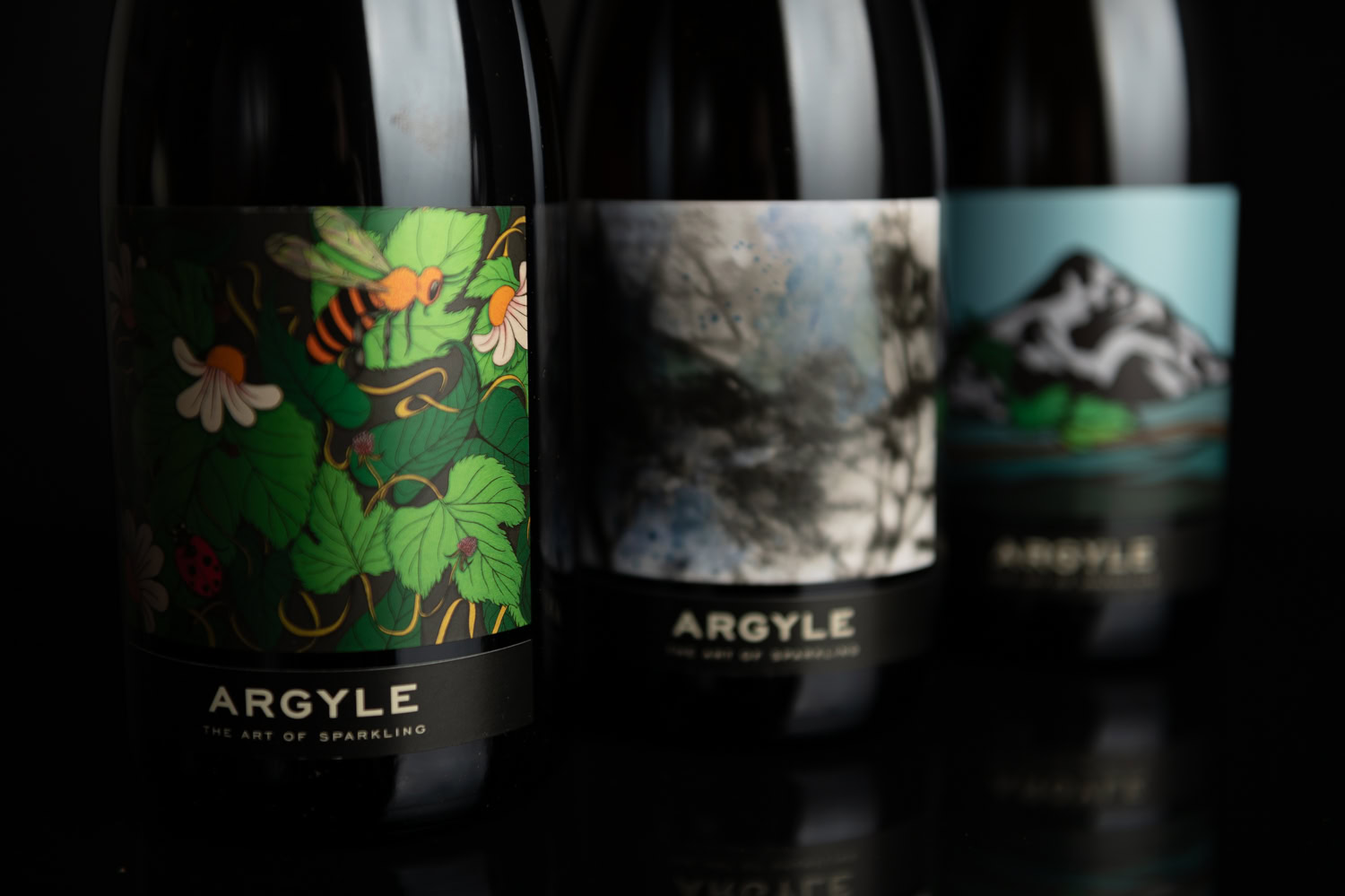 Two wine bottles are shown in a dark setting. The first bottle features an illustrated label with a bee and leaves, the second displays an abstract cloudy design. The labels all read "ARGYLE" with the tagline "The Art of Sparkling."