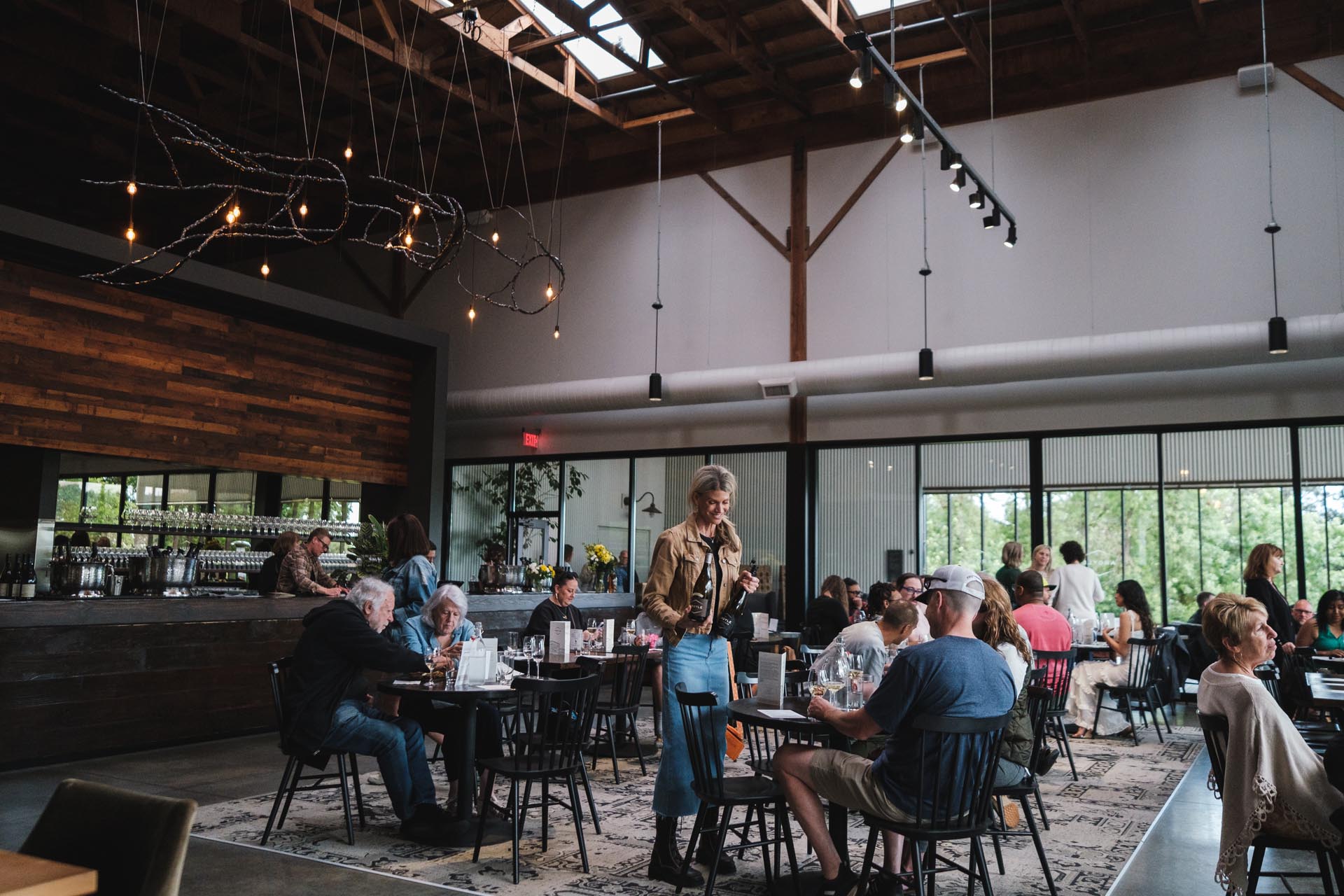 A bustling Argyle Winery Tasting Room with wooden accents and an open ceiling. Guests enjoy their wines while a Tasting Room server in a brown jacket attends to guests. The bar area is visible on the left, and a large window on the right lets in natural light.