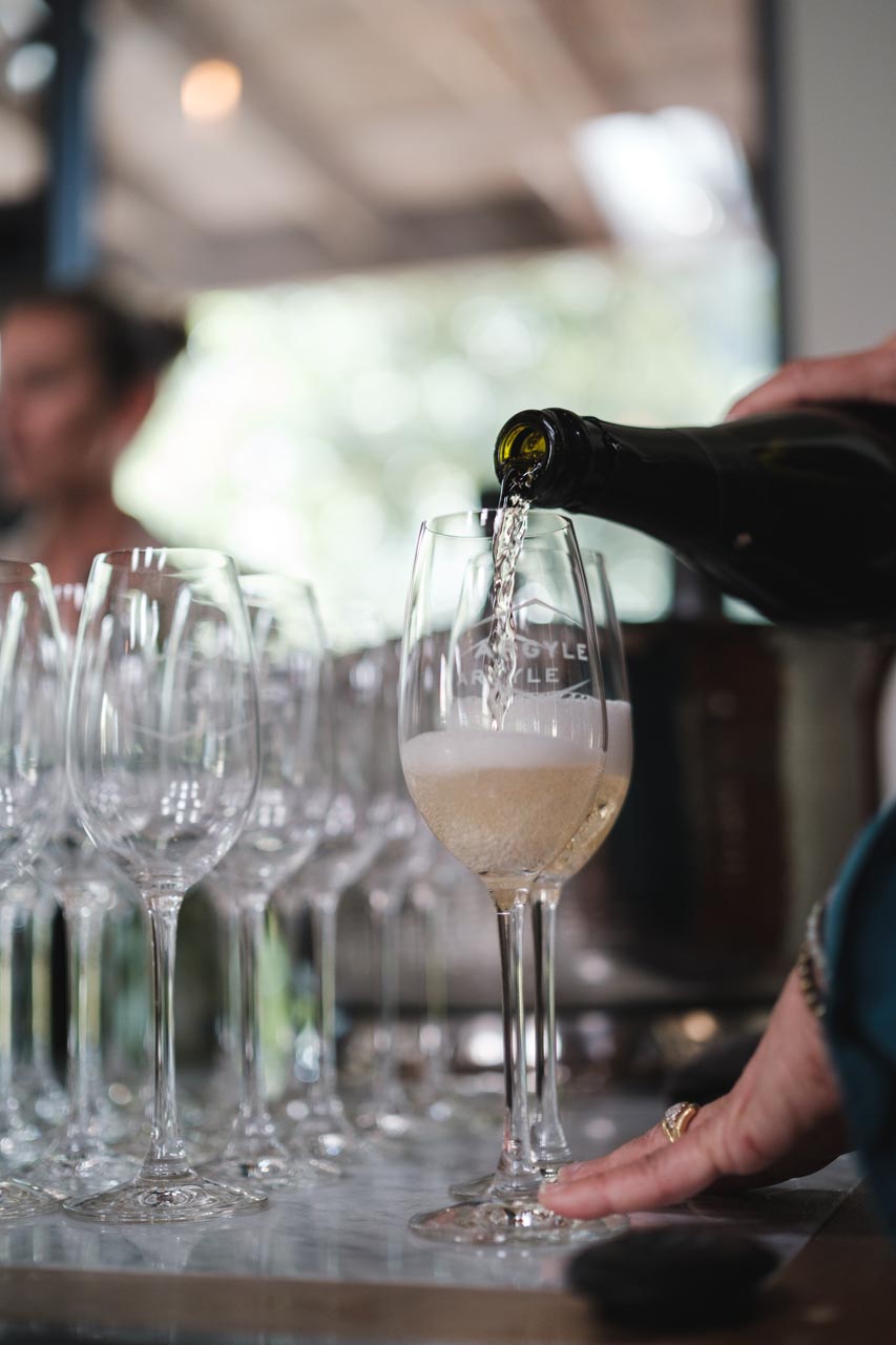 A hand is pouring Argyle sparkling wine into a glass from a dark bottle. Several empty wine glasses are arranged nearby. The blurred background suggests an indoor setting with natural light. The scene captures a moment of wine service.