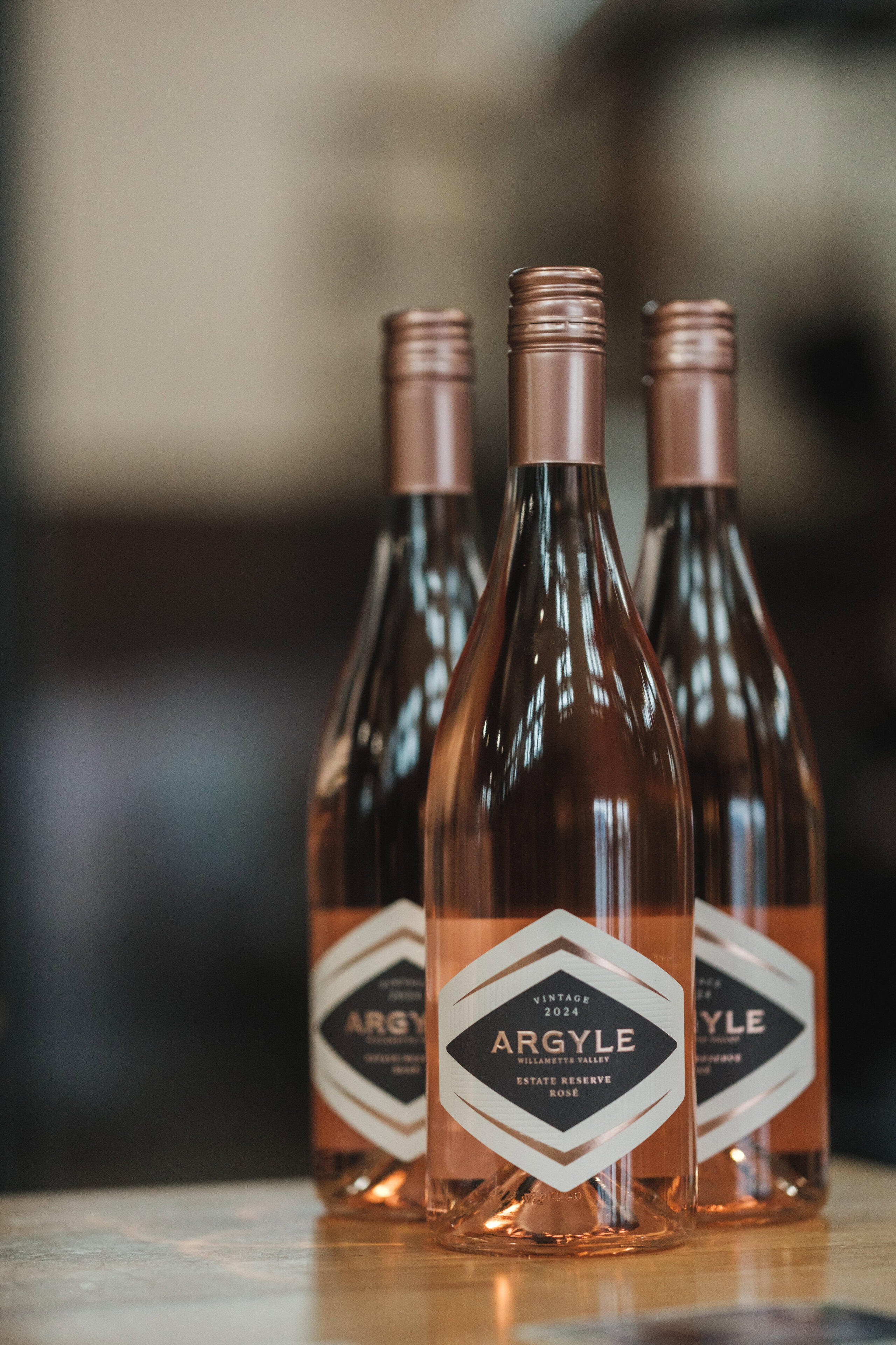 Three bottles of Argyle 2024 Estate Reserve Rosé standing on a wooden surface, with soft indoor lighting reflecting off the glass.