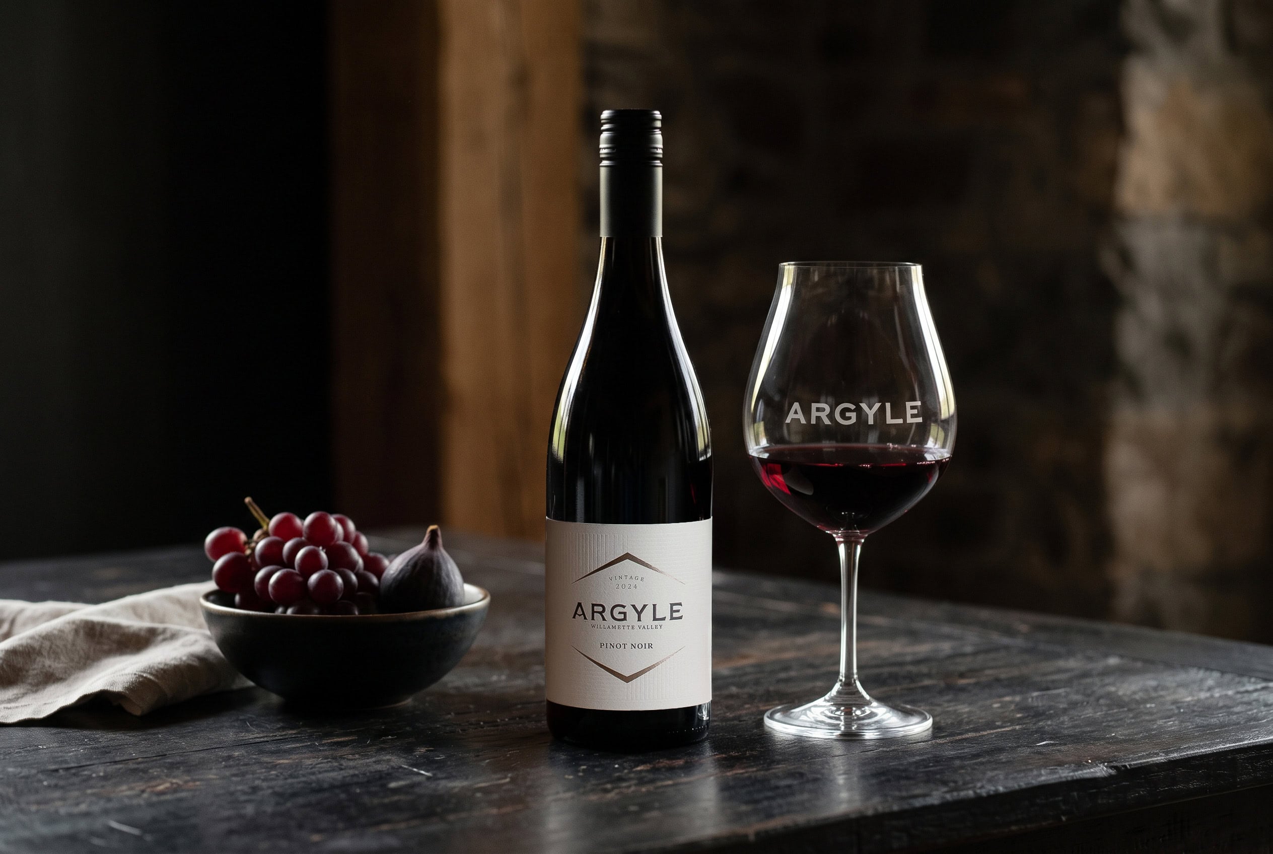 Argyle 2024 Willamette Valley Pinot Noir beside an etched Argyle glass and a bowl of red grapes and figs on a dark wood table