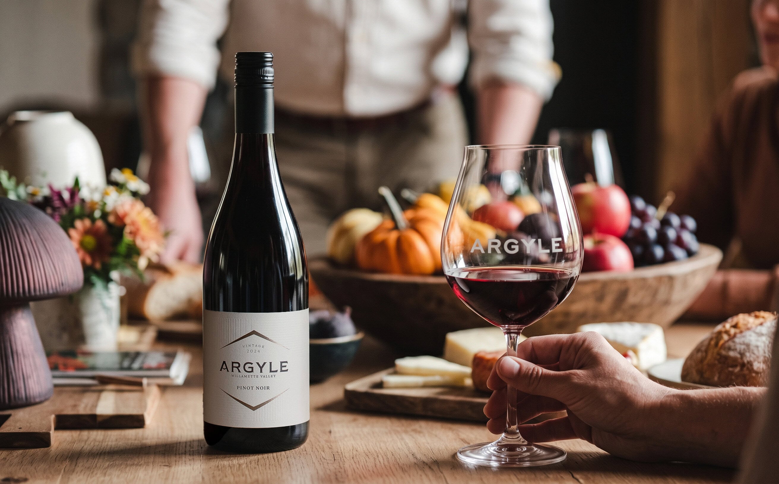 A bottle of Argyle 2024 Willamette Valley Pinot Noir on a harvest table with pumpkins, apples, cheese and a filled Argyle glass