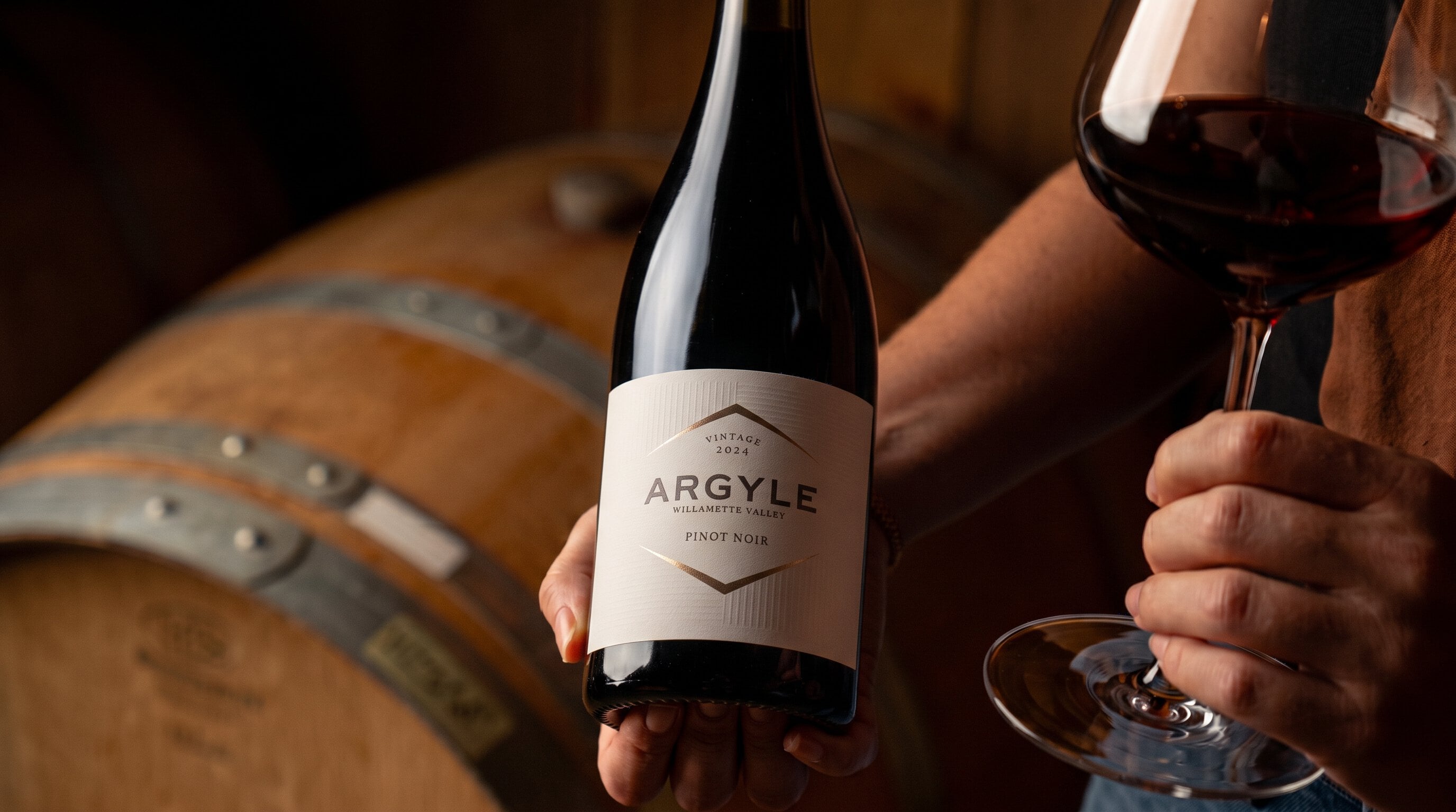 A hand holding a bottle of Argyle 2024 Willamette Valley Pinot Noir and a glass of red wine beside oak barrels in the cellar